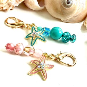 May include: Two gold-toned keychains with decorative charms. One features a pink and gold starfish charm, the other a turquoise starfish. Both have colorful beads in shades of pink, turquoise, and blue. Seashells are in the background.