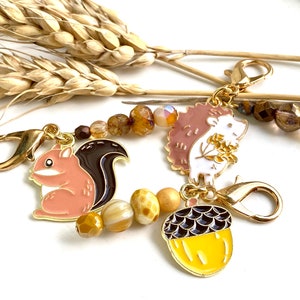 Könnte beinhalten: Ein goldfarbenes Armband mit einem Eichhörnchen-, Igel- und Eichel-Anhänger. Die Anhänger sind emailliert und haben ein detailliertes Design. Das Armband besteht aus braunen und goldenen Perlen.
