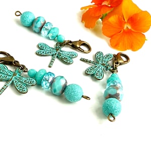 May include: Close-up of several teal-colored beaded charms with dragonfly pendants and bronze-colored clasps. The beads vary in size and texture, with some having a frosted appearance. An orange flower is in the background.