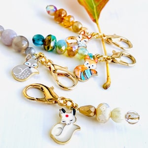 May include: A set of four fox-themed stitch markers with gold-toned metal clasps and colourful glass beads. The markers feature different fox designs, including a white fox, a grey fox, a red fox, and a fox with a blue and green patterned tail.