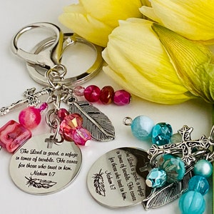 May include: Two keychains with religious inscriptions and decorative beads. Each keychain features a round, silver-colored charm with the text "The Lord is good, a refuge in times of trouble. He cares for those who trust in him. Nahum 1:7."