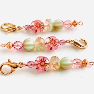 May include: Three gold-toned beaded charms with pink and green glass beads. Each charm features a pink flower bead and a green striped bead. The charms have a gold-toned lobster clasp.