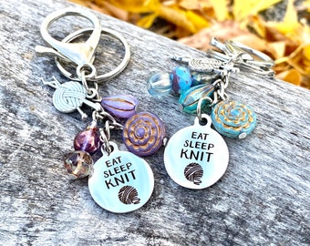 May include: A silver keychain with a charm that says "Eat Sleep Knit" and a small yarn ball charm. The keychain also has several colorful beads, including purple, blue, and gold.