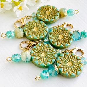 May include: Five teal green and gold patterned round charms with gold clasps and aqua blue glass beads. The charms have a geometric design.