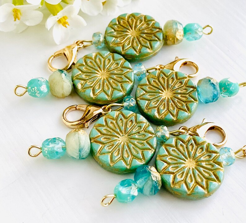 May include: Five teal green and gold patterned round charms with gold clasps and aqua blue glass beads. The charms have a geometric design.