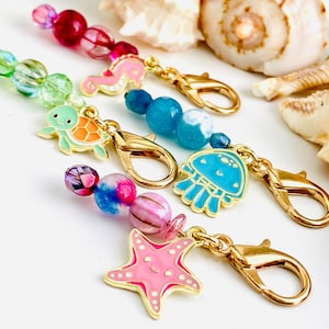 May include: A set of five colorful charm necklaces with different sea-themed charms, including a turtle, a starfish, a jellyfish, a seahorse, and a beaded charm. Each charm is attached to a gold chain with a lobster clasp.