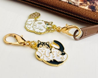 Op de afbeelding: Twee gouden sleutelhangers met kattencharms. De ene sleutelhanger heeft een witte kat met zwarte vlekken en een zwarte staart. De andere sleutelhanger heeft een zwarte kat met witte vlekken en een witte staart.