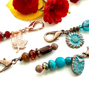 May include: Four metal lobster clasp charms featuring decorative beads and pendants. One charm has red beads and a maple leaf, one has blue beads and a sunflower, one has brown beads, and one has turquoise beads with an owl pendant.