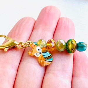 May include: A gold charm featuring a fox design and colourful beads. The fox is orange and white with blue and green accents. The beads are amber, green, and blue.