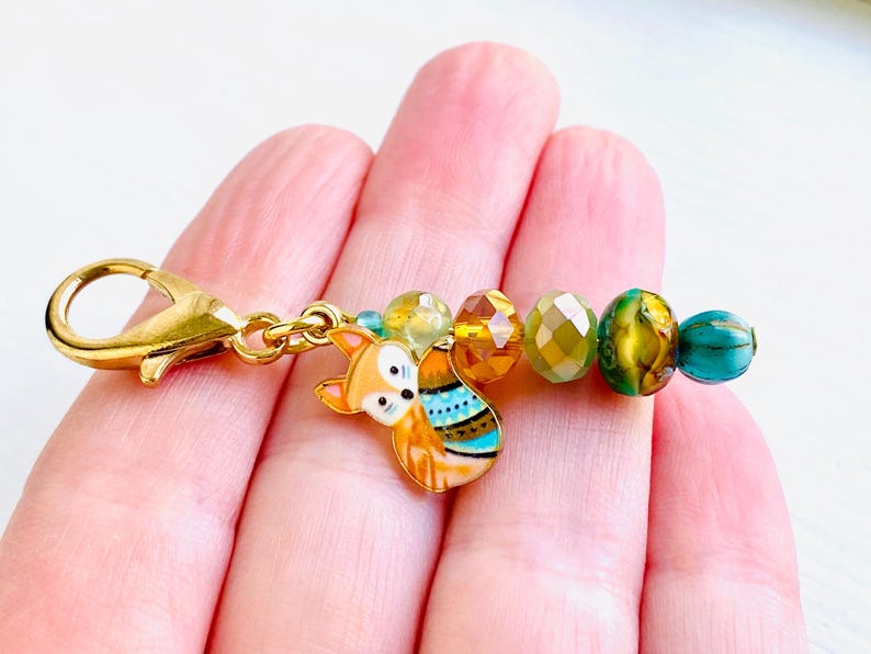 May include: A gold charm featuring a fox design and colourful beads. The fox is orange and white with blue and green accents. The beads are amber, green, and blue.