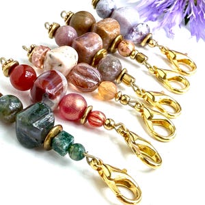 May include: A selection of beaded keychains with gold-coloured clasps. The beads are in various shapes and colours, including red, green, brown, and purple. The keychains are arranged diagonally against a white background, with a purple flower in the upper right corner.