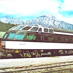 May include: Vintage postcard featuring a CN train with a distinctive observation car. The train is white and black, with a glass-domed roof, set against a backdrop of mountains and a blue sky. The postcard is a vintage item.