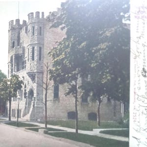 May include: Vintage postcard featuring a stone castle-like building with multiple turrets and arched windows. A tree-lined street leads to the building, with a statue visible in the distance. The postcard has a faded, aged appearance.