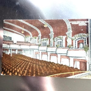 May include: Vintage postcard depicting the interior of the Orpheum Theatre in Quincy, IL. The image shows rows of brown seats facing a stage, with balconies and decorative architectural details. The ceiling is arched and painted in shades of red and white.