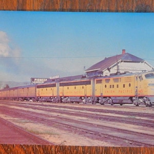 May include: A yellow and blue Union Pacific train with a steam engine pulling a long line of passenger cars. The train is traveling through a small town with a building in the background. The sign "Pioneer Lumber" is visible on the side of the building.