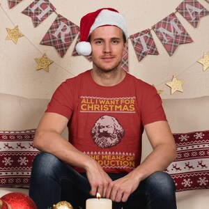 以下が含まれることがあります： サンタの帽子をかぶった人物が赤いTシャツを着ています。Tシャツにはカール・マルクスの肖像画と「クリスマスに欲しいものは生産手段だけ」という文字が書かれています。