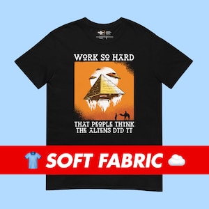 Puede incluir: Camiseta negra con un diseño gráfico de una pirámide con un fondo de puesta de sol. El texto "WORK SO HARD THAT PEOPLE THINK THE ALIENS DID IT" está impreso en la camiseta.