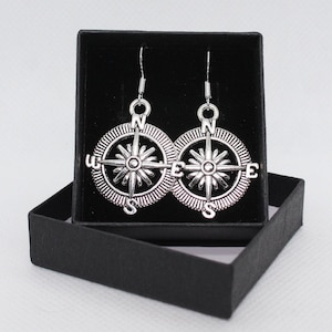 Puede incluir: Un par de pendientes de plata en forma de brújula en una caja de regalo negra. Los pendientes presentan un diseño de rosa de los vientos con las letras N, S, E y W.