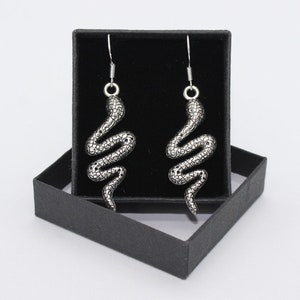 May include: A pair of silver snake earrings in a black jewelry box. The earrings are detailed with a textured snake design.