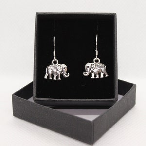 Puede incluir: Un par de pendientes de elefante colgantes de plata en una caja de regalo negra. Los pendientes presentan diseños detallados de elefantes con superficies texturizadas.