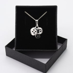 May include: A silver necklace with a charm of three stacked buttons. The necklace is displayed in a black velvet box.