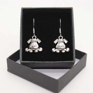 May include: A pair of silver dangle earrings featuring a cartoon dog design. The earrings are displayed in a black velvet box.