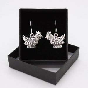 May include: A pair of silver chicken earrings in a black gift box. The earrings are detailed and have a rustic look.