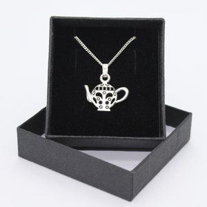 May include: A silver teapot pendant necklace in a black velvet box. The teapot has a black and white patterned design.