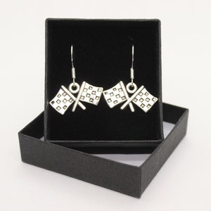 May include: Silver-coloured earrings with a chequered flag design, displayed in a black gift box. The earrings have a hook fastening and a detailed chequered pattern. The box is open, revealing the jewellery against a dark background.