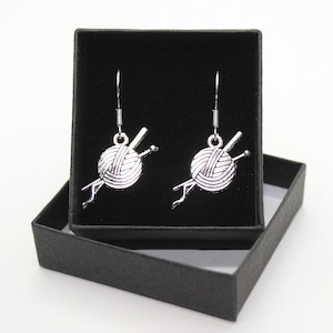 May include: A pair of silver earrings with a yarn ball and knitting needles charm. The earrings are displayed in a black velvet box.