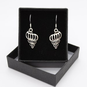 May include: Silver seashell earrings presented in a black jewellery box. The earrings feature an intricate, open design, suspended from simple ear wires. The box is open, showing a black velvet interior.