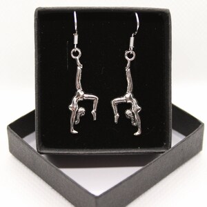 May include: A pair of silver earrings featuring a gymnast in a handstand pose. The earrings are displayed in a black box.