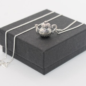 Puede incluir: Un collar de plata con un dije de tetera. La tetera tiene un diseño detallado y las palabras "tea time" grabadas en ella. El collar se muestra en una caja negra.