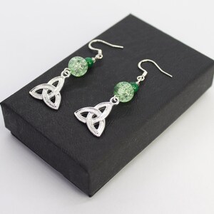 May include: Silver Celtic knot earrings with green glass beads. The earrings are on a black background.