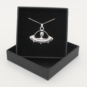 May include: A silver pendant necklace in a black velvet box. The pendant is a silver UFO with an alien inside and the words "AWAIT TO BELIEVE" inscribed on the side.
