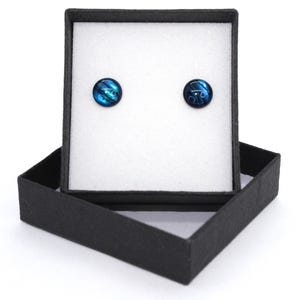 May include: A pair of round stud earrings featuring a deep blue and black iridescent pattern. The earrings are displayed on a white foam insert inside a square black cardboard gift box with the lid propped open behind the base.