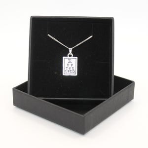Puede incluir: Un collar plateado con un colgante con forma de tabla optométrica, presentado en una caja de regalo negra. El colgante tiene las letras E, FP, TOZ y LPED. El collar se muestra sobre un fondo blanco.