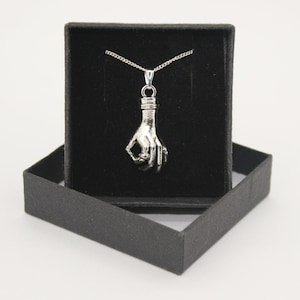 Puede incluir: Un collar de plata con un colgante con forma de mano haciendo el signo de OK. El collar está en una caja de terciopelo negro.