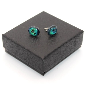 May include: A pair of oval stud earrings featuring a smooth, glossy surface with swirling patterns of deep blue, teal, and black. The earrings are displayed on a square, black textured jewellery box with a matte finish and a hinged lid design.