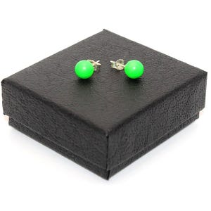 May include: A pair of bright neon green stud earrings featuring spherical beads attached to silver-toned metal posts. The earrings are displayed on top of a square, black textured jewelry box with a lid that has a slightly raised edge.