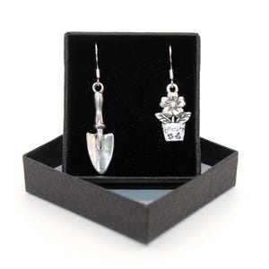 Puede incluir: Pendientes colgantes plateados, uno con forma de pequeña paleta de jardín y el otro con una flor en una maceta. El pendiente de maceta tiene la palabra "Nature". Los pendientes se muestran en una caja de regalo negra.