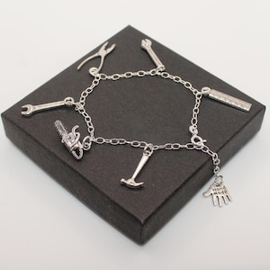 Puede incluir: Pulsera de plata con una variedad de dijes de herramientas, incluyendo una llave, un martillo, una sierra y una regla. La pulsera también tiene un dije de mano con las palabras "Hand Made" en él.