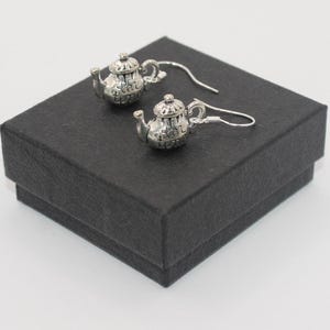 Puede incluir: Un par de pendientes de plata en forma de tetera en una caja de regalo negra. Las teteras tienen un diseño detallado y las palabras "Me encanta el té" están inscritas en ellas.