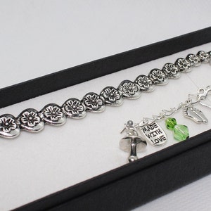 May include: Silver bookmark with a chain of linked hearts, a charm with the words "Made with Love", a green crystal, and a baby bottle charm. The bookmark is in a black gift box.