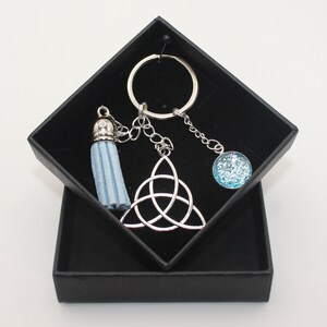 May include: A silver key chain with a blue tassel, a silver Celtic knot charm, and a blue glitter charm. The key chain is in a black gift box.