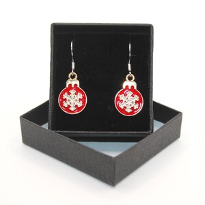 May include: A pair of Christmas earrings. Each earring has a red, round ornament design with a silver snowflake and a silver hook. The earrings are presented in a black gift box.