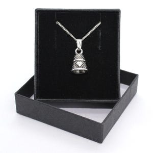 Puede incluir: Un collar con colgante en forma de dedal plateado que presenta una superficie texturizada y un pequeño detalle de corazón en la parte frontal. El colgante cuelga de una delicada cadena plateada, presentado en un joyero cuadrado de color negro.