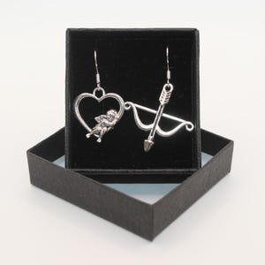 May include: A pair of silver earrings in a black box. One earring features a heart with a cherub inside, the other earring features a bow and arrow.