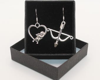 Puede incluir: Un par de pendientes de plata en una caja negra. Un pendiente tiene un corazón con un querubín dentro, el otro pendiente tiene un arco y una flecha.