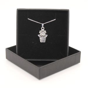 Puede incluir: Un collar plateado con un colgante en forma de flor en maceta, presentado en una caja de regalo negra. El colgante tiene la palabra "Nature" grabada. El collar se muestra sobre un fondo blanco.